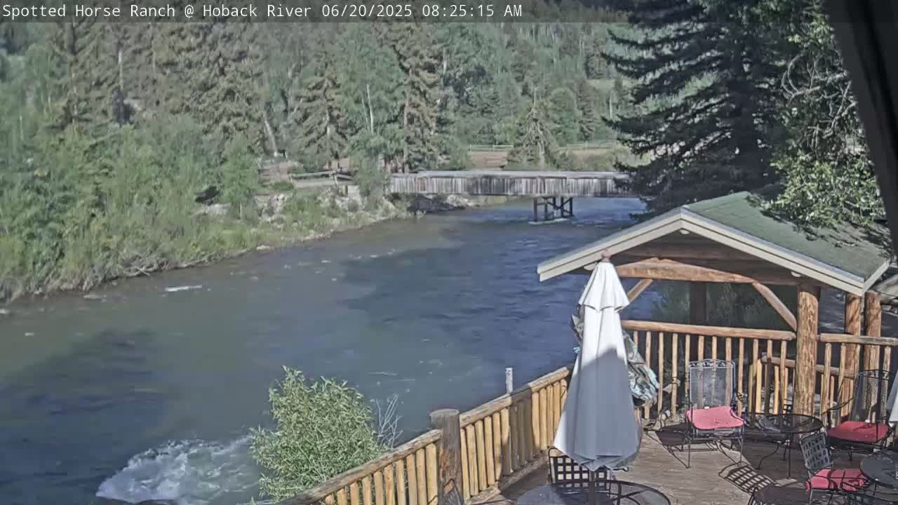 A deck with wooden railings and outdoor furniture overlooks a flowing river under a sunny sky, with a wooden bridge visible in the distance.