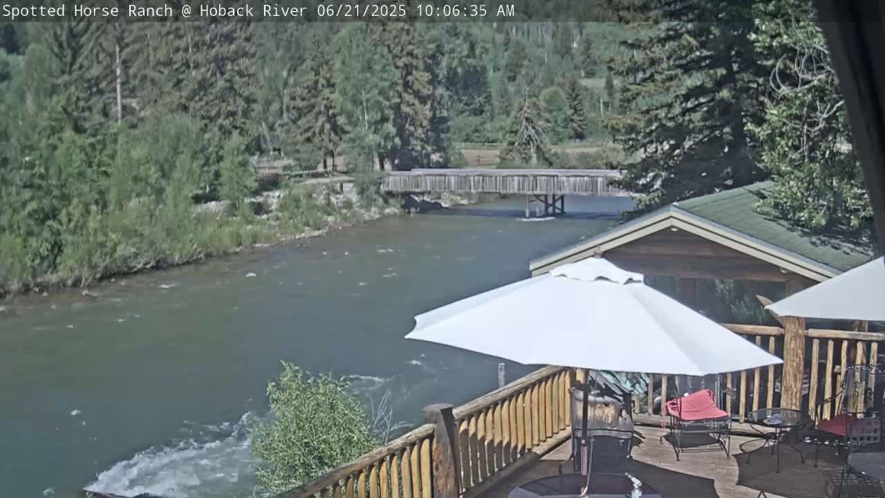 A deck with a wooden railing overlooks a river flowing under a wooden bridge, with a white umbrella providing shade on a partly sunny day.