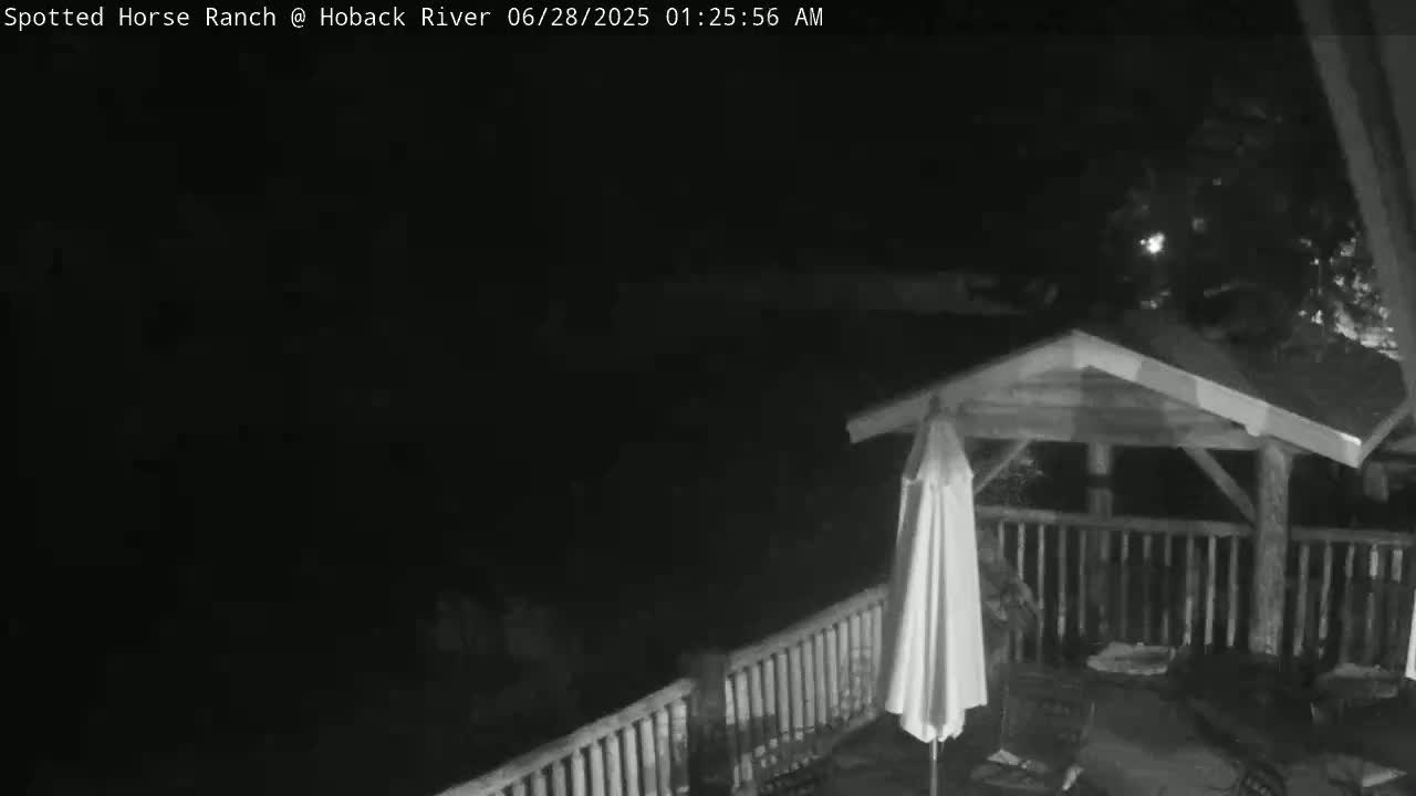 A nighttime, low-light image shows a wooden gazebo with a patio set and a closed patio umbrella on a deck with railings.