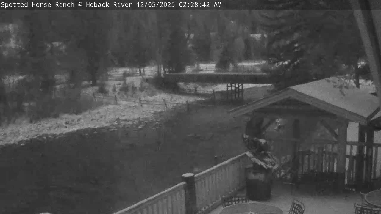 The dark, snowy scene depicts a river flowing past a snow-dusted bank, with a distant bridge and trees, and a foreground patio featuring outdoor furniture and a covered structure, all under cold, early morning conditions.