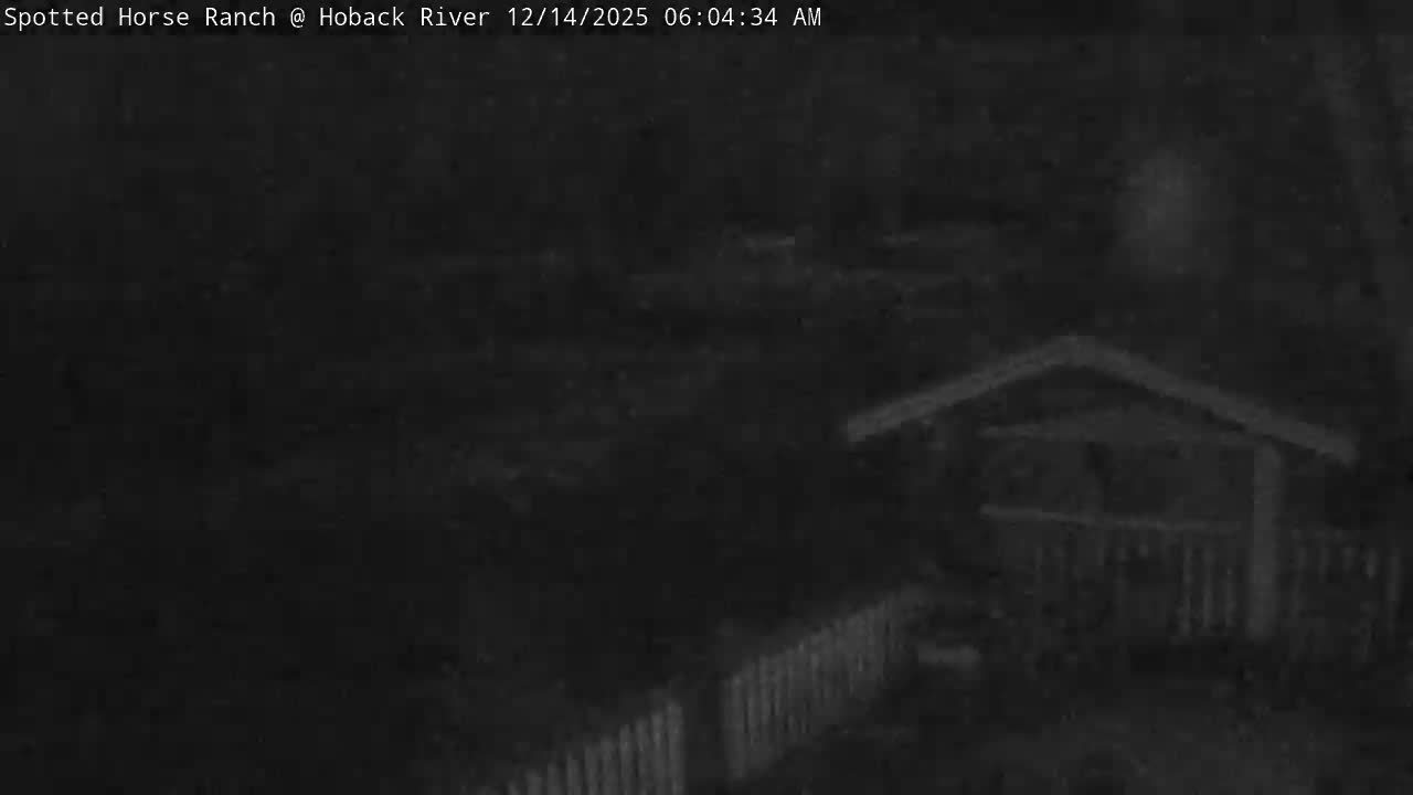 The dark, snowy scene depicts a river flowing past a snow-dusted bank, with a distant bridge and trees, and a foreground patio featuring outdoor furniture and a covered structure, all under cold, early morning conditions.