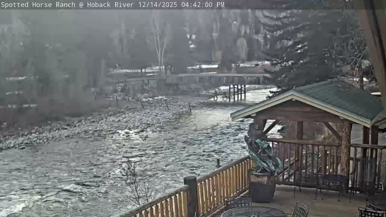 The dark, snowy scene depicts a river flowing past a snow-dusted bank, with a distant bridge and trees, and a foreground patio featuring outdoor furniture and a covered structure, all under cold, early morning conditions.