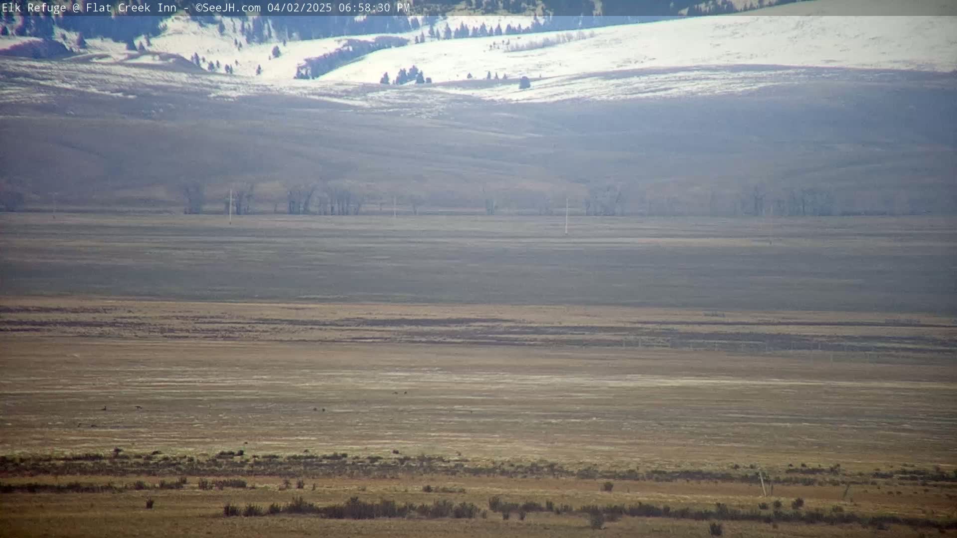 A vast, flat expanse of dry, brown land stretches to snow-covered hills in the distance under a hazy sky.