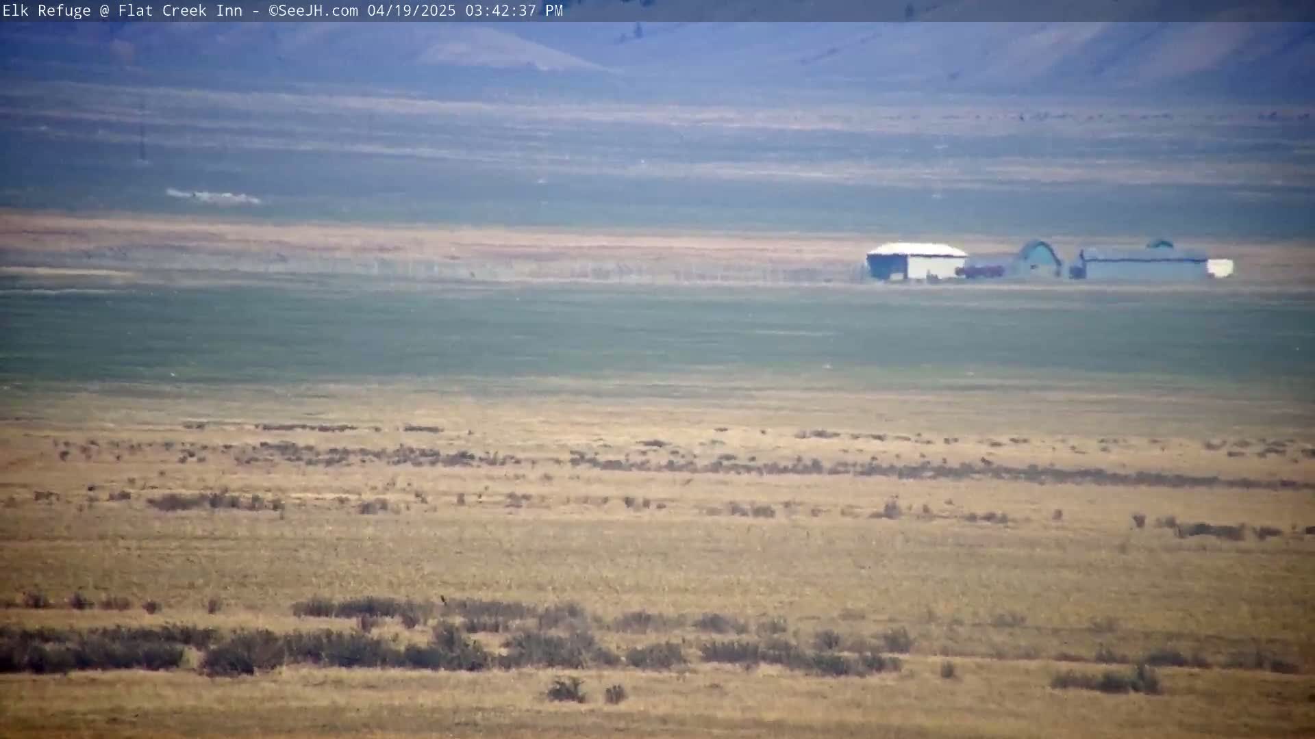 A hazy, long-distance view of a flat, dry landscape with sparse vegetation and a few buildings in the mid-ground.