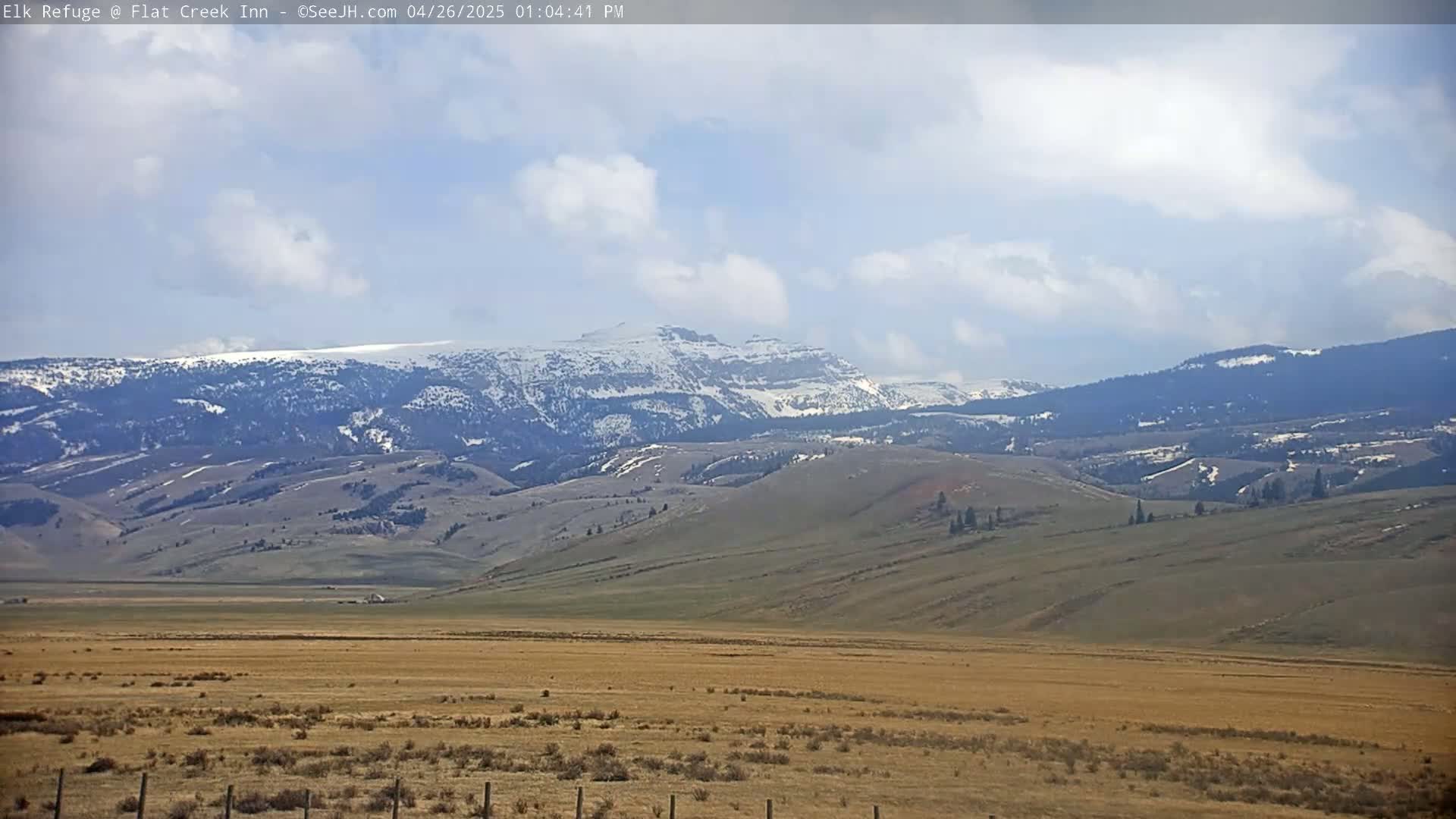 A partly cloudy day reveals a vast, tan field extending to snow-capped mountains in the distance.