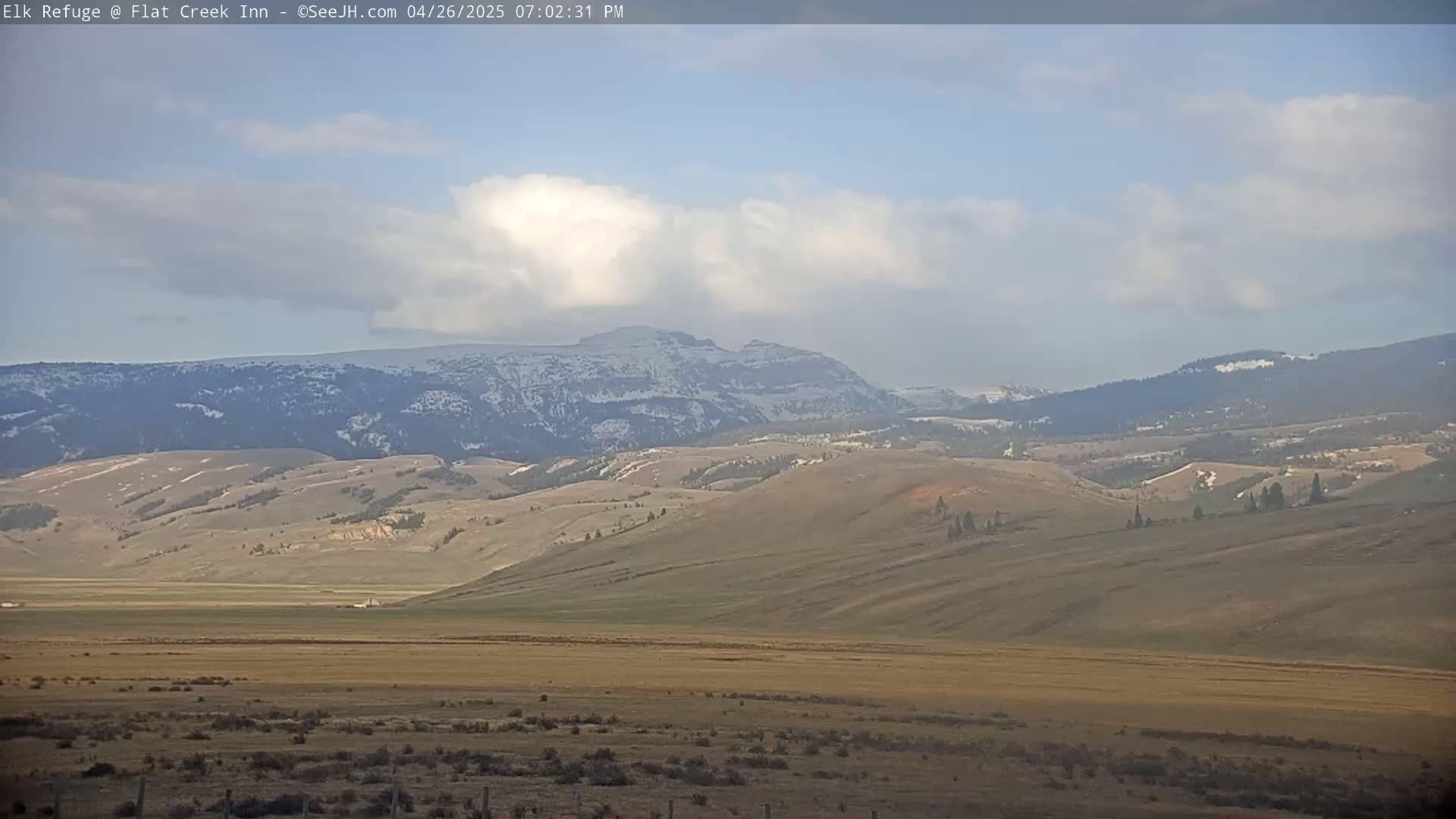 A mostly sunny, partly cloudy day reveals a vast expanse of rolling hills and mountains, the latter dusted with snow.