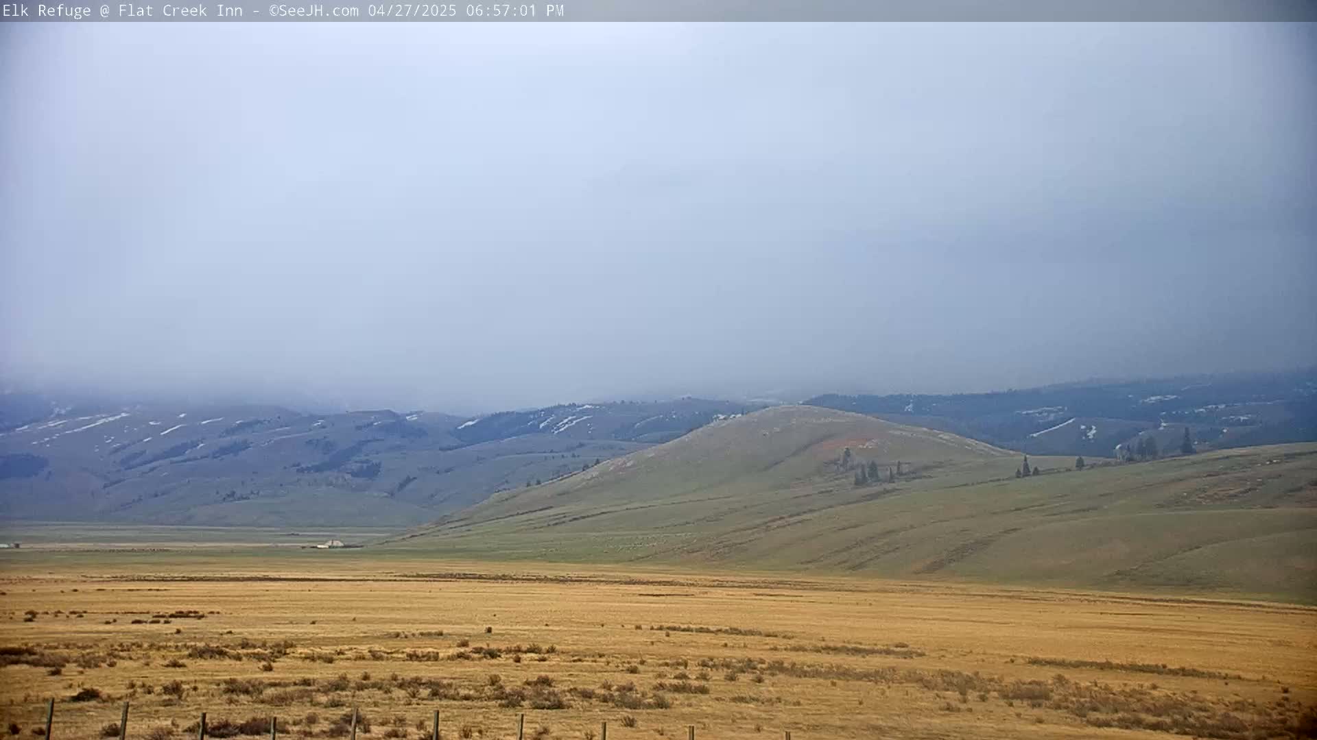 A hazy view shows a vast, dry, tan plain stretching to rolling green hills and distant mountains with patches of snow under a light gray sky.