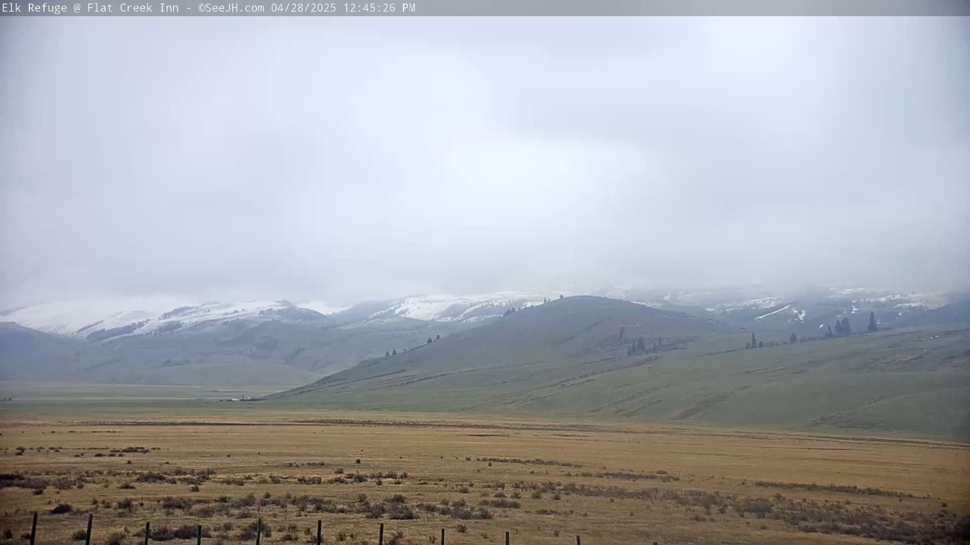 A hazy, flat valley stretches to snow-dusted mountains under a mostly cloudy sky.