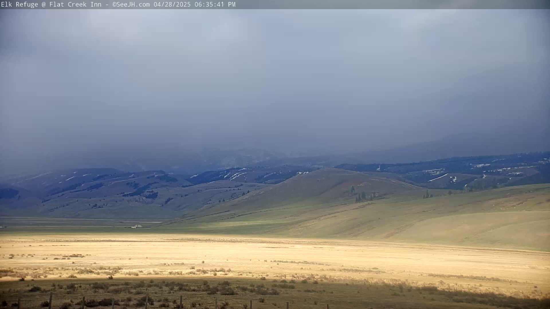 A hazy, sunlit landscape reveals rolling green and tan hills under a muted, overcast sky.
