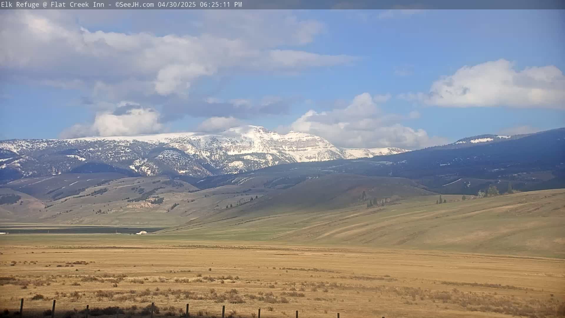 A partly cloudy blue sky overlooks a vast, tan plain stretching to snow-capped mountains in the distance.