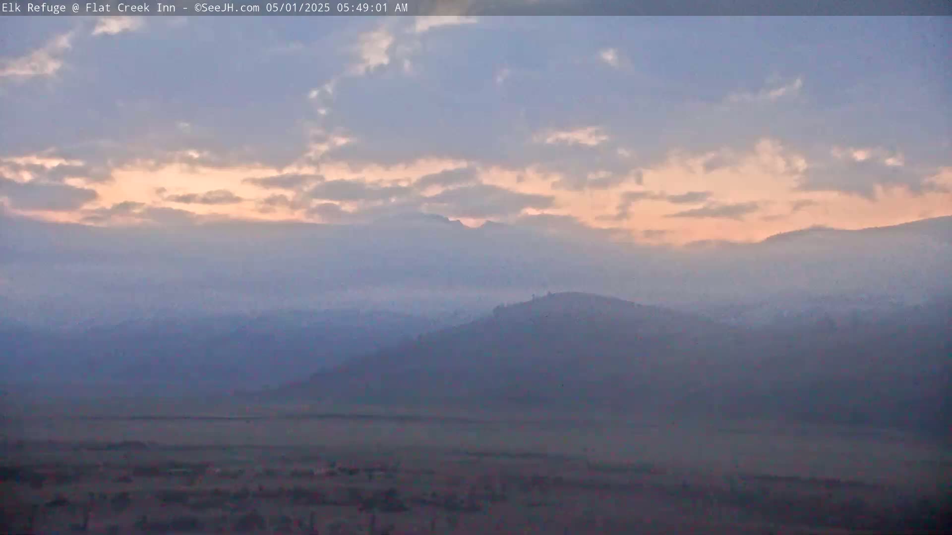 A hazy sunrise illuminates a landscape of rolling hills and distant mountains under a partly cloudy sky.