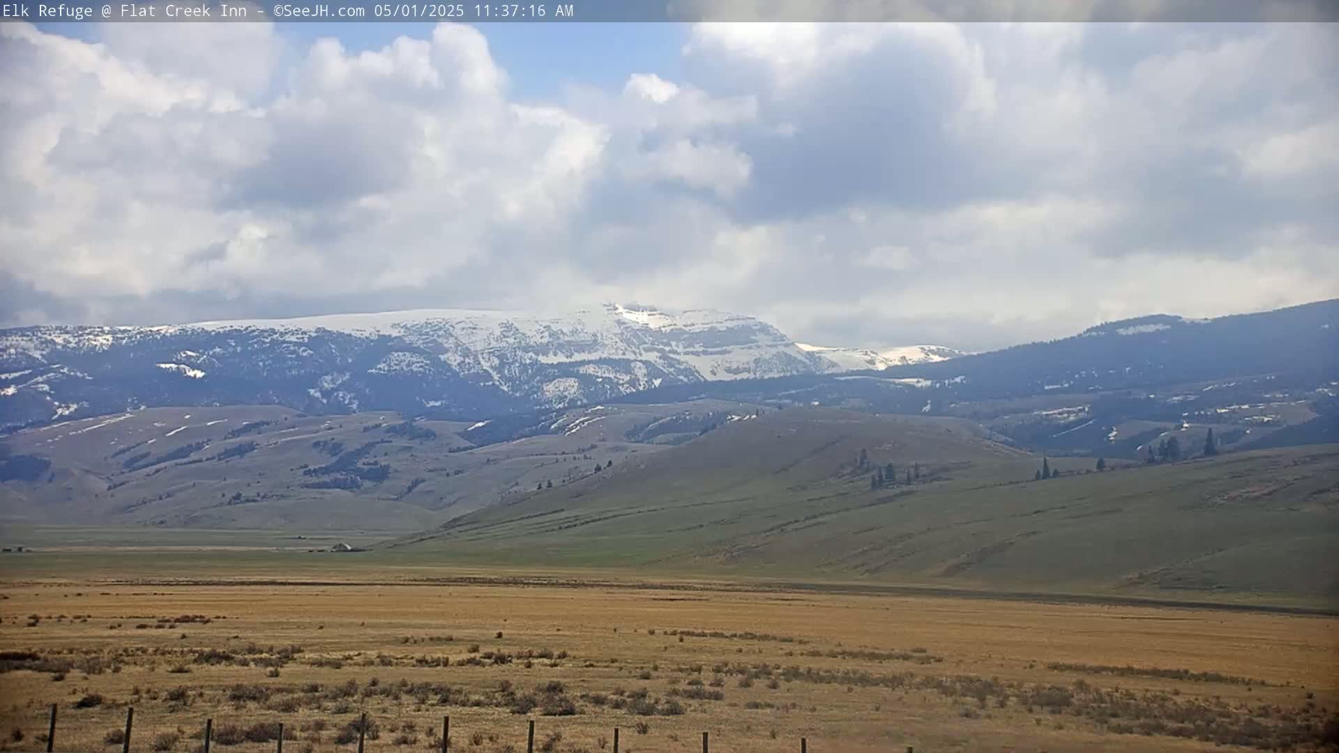 A partly cloudy day reveals a vast, rolling landscape of tan and green hills leading to snow-capped mountains in the distance.