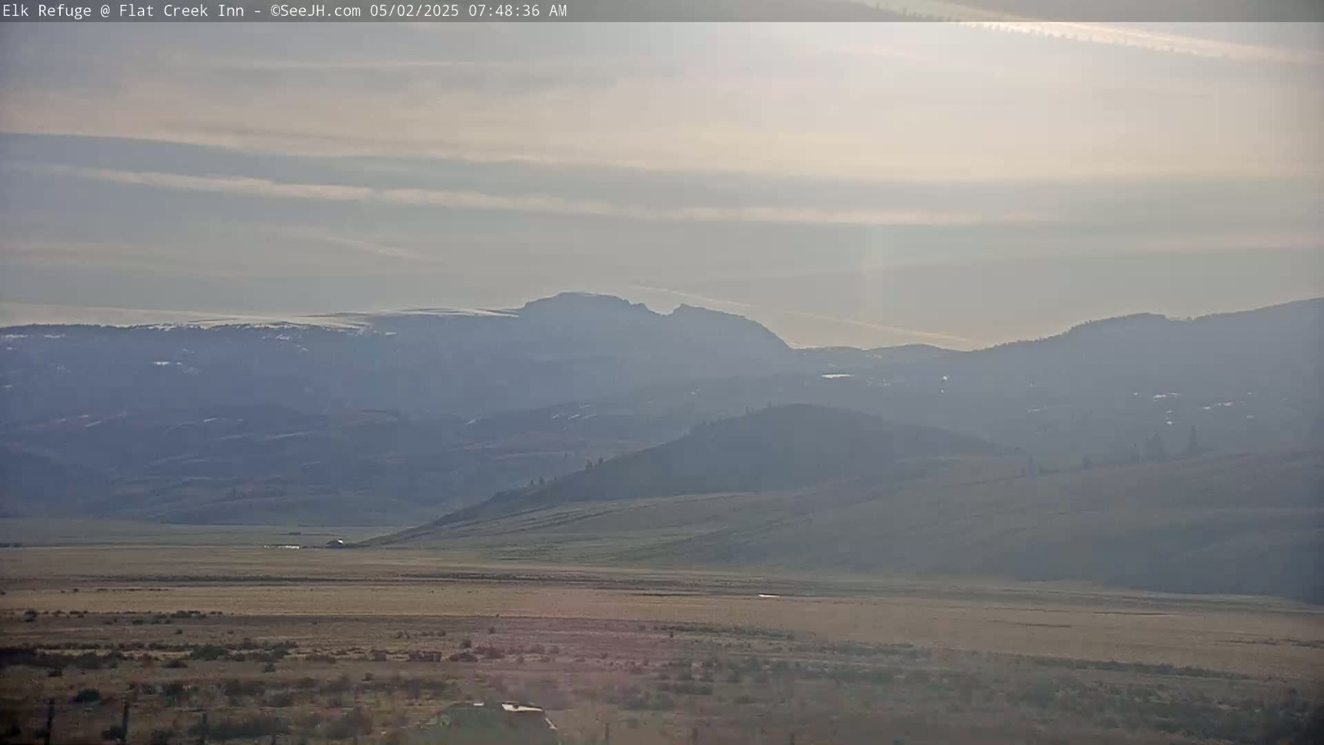 A hazy, sunlit landscape shows a flat, dry valley stretching to distant, snow-capped mountains under a pale sky.