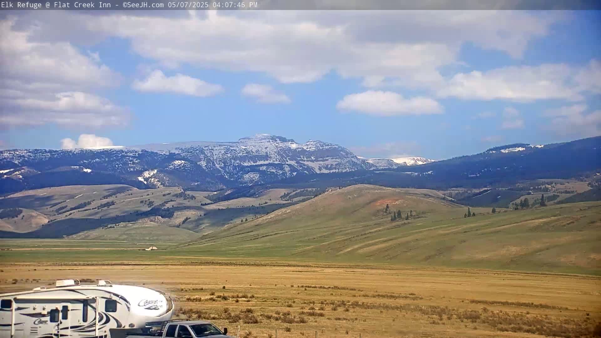 A partly cloudy sky overlooks a vast, rolling landscape of tan and green hills leading to snow-capped mountains in the distance, with a recreational vehicle and truck parked in the foreground.