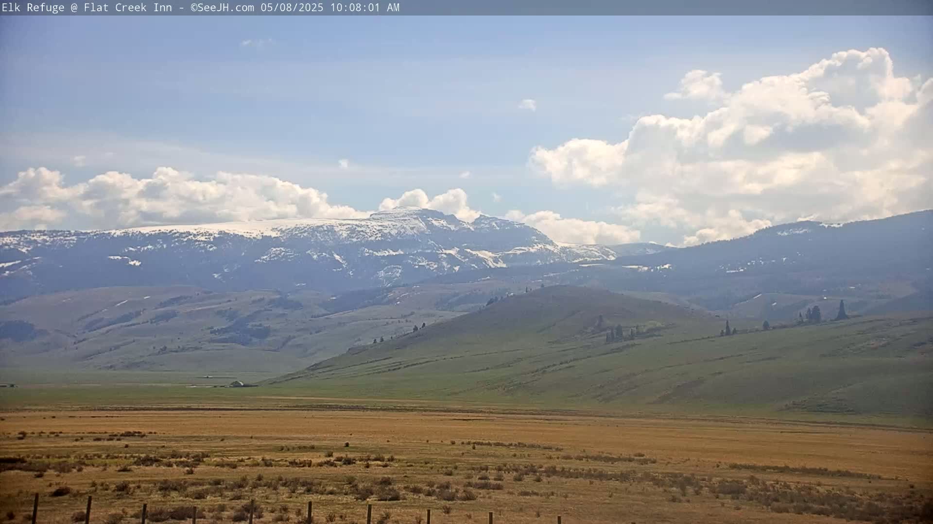 A partly cloudy day reveals snow-capped mountains in the distance, overlooking a vast expanse of rolling hills and plains.