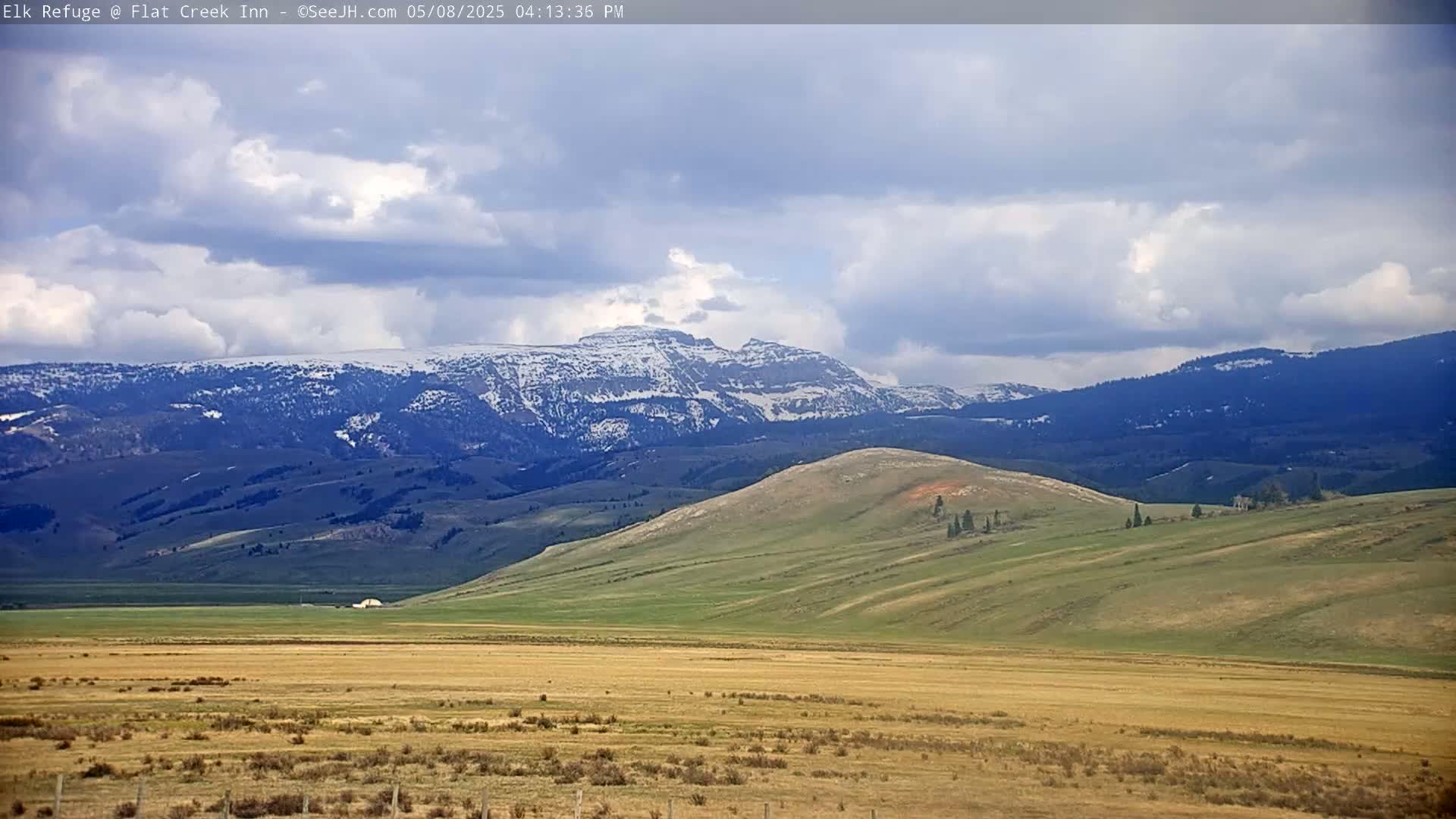 A partly cloudy sky overlooks a vast, rolling landscape of green and tan hills and plains, with snow-capped mountains in the background.