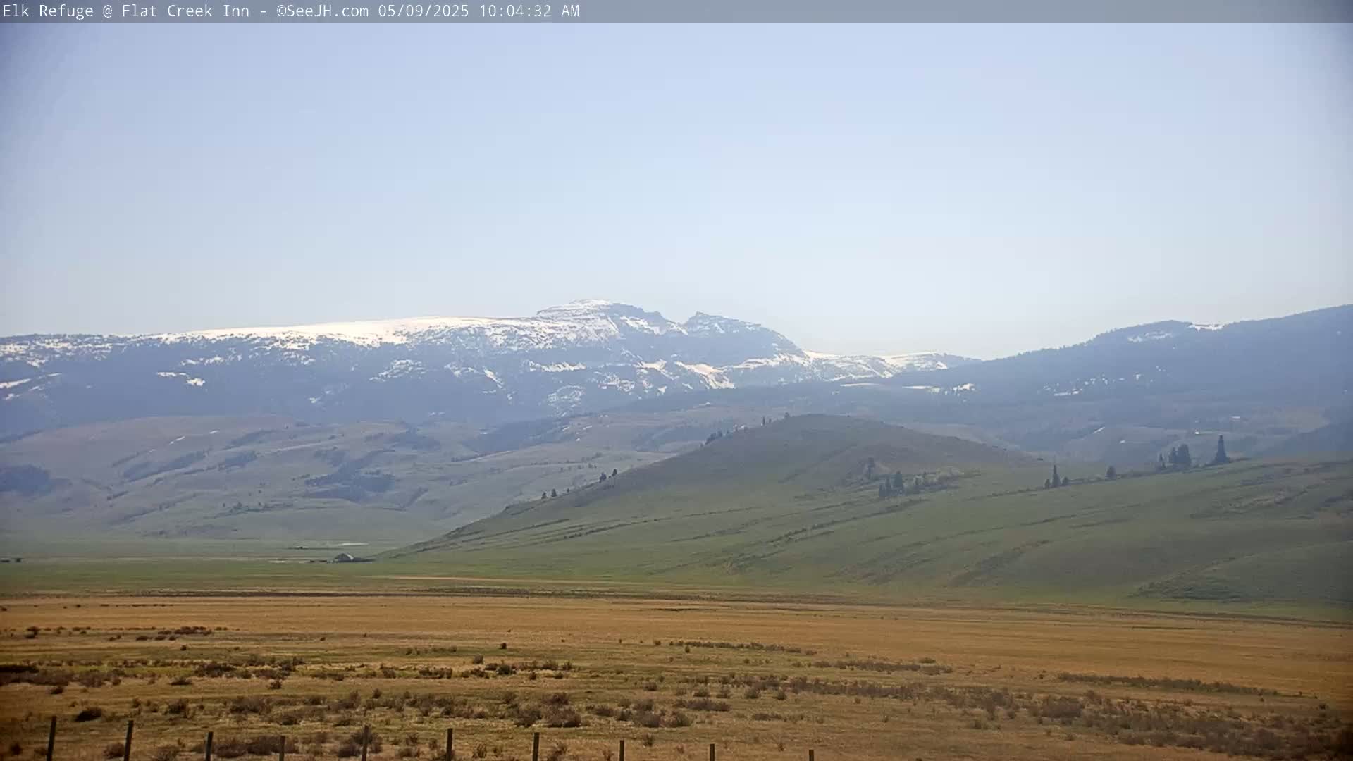 A long-range view shows rolling hills and distant mountains, the highest of which have patches of snow, under a clear, light-blue sky.