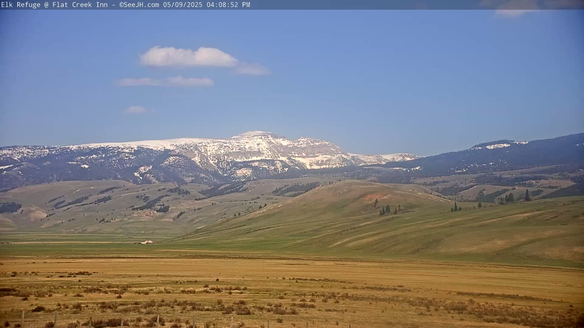 A snow-capped mountain range is visible in the distance under a mostly clear blue sky, overlooking a vast expanse of tan and green rolling hills and plains.