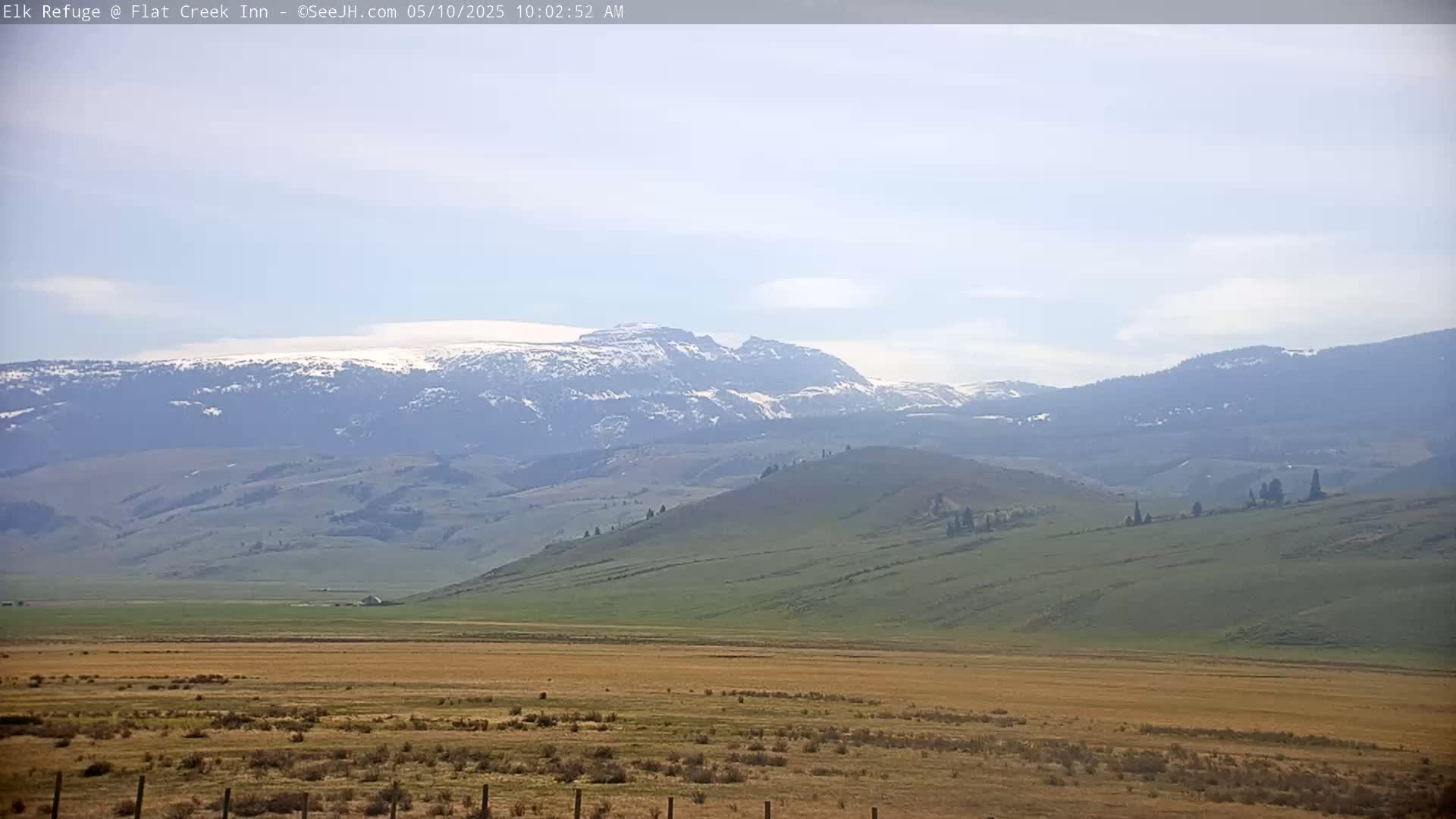 A mostly sunny day reveals a vast, flat, dry field leading to rolling green hills and a distant mountain range with snow-capped peaks.
