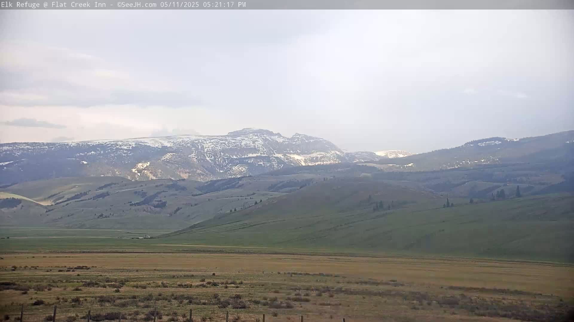 A hazy view shows a vast, flat valley of brown and green grass leading to rolling, green hills and distant mountains partially capped with snow.