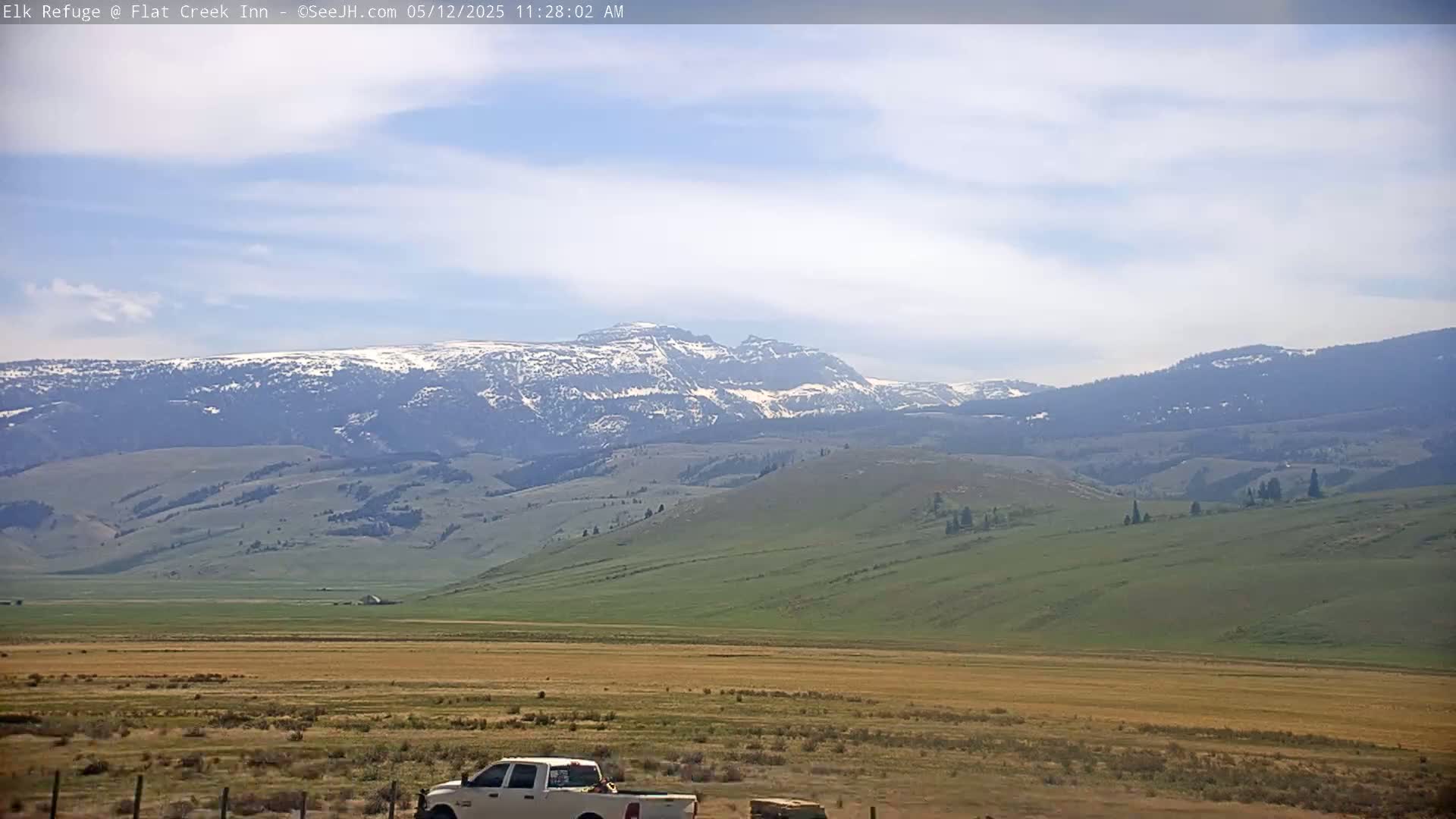 A snow-capped mountain range is visible in the distance under a partly cloudy sky, with a flat, grassy plain and a white pickup truck in the foreground.