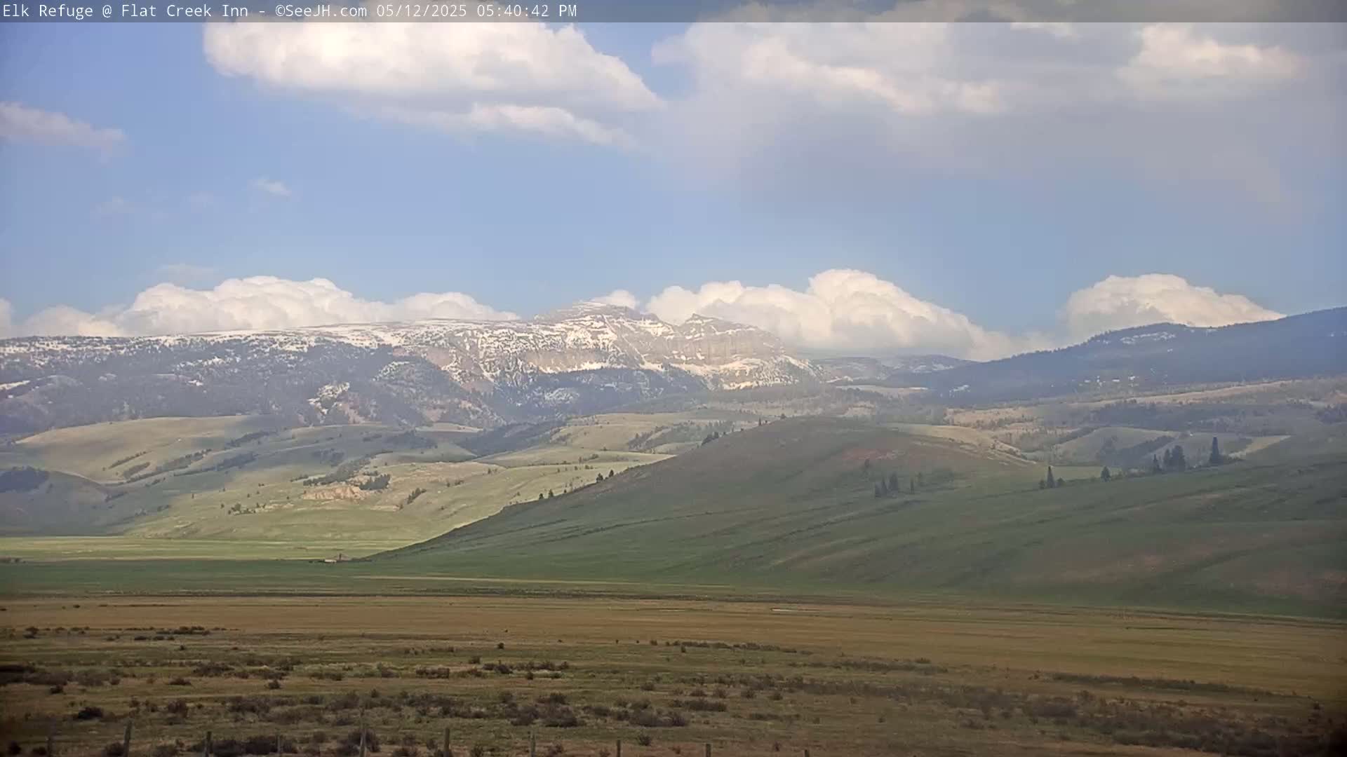 Rolling green hills and mountains with patches of snow under a partly cloudy sky.