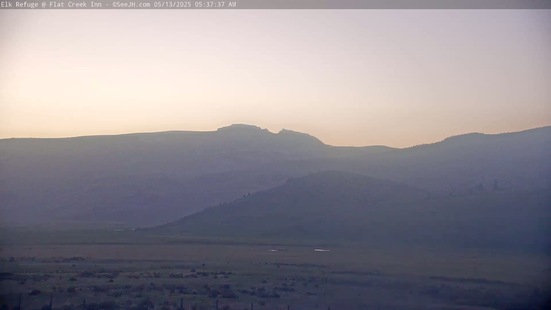 A hazy sunrise illuminates a flat valley and rolling hills, with a prominent, distant mountain range silhouetted against the pale sky.