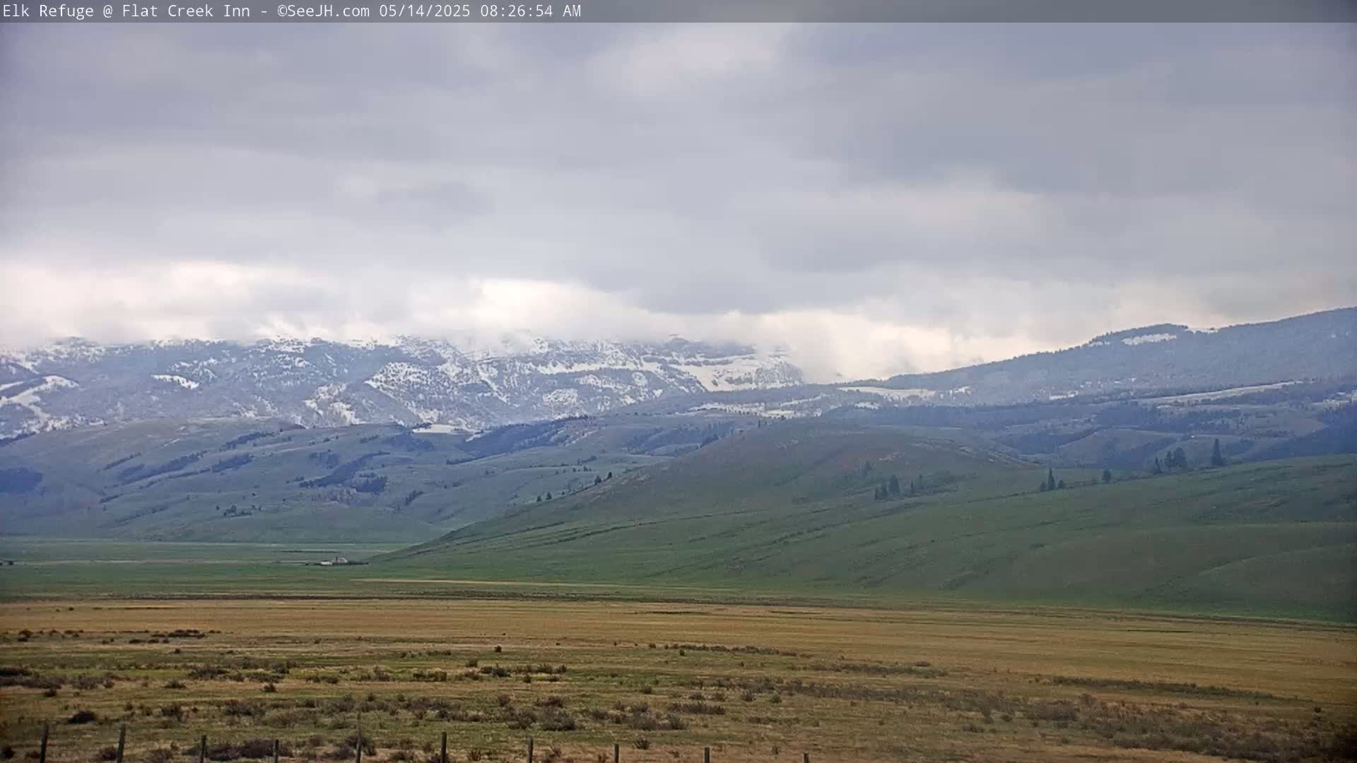 Snow-capped mountains rise in the distance behind a flat, grassy valley under a cloudy sky.