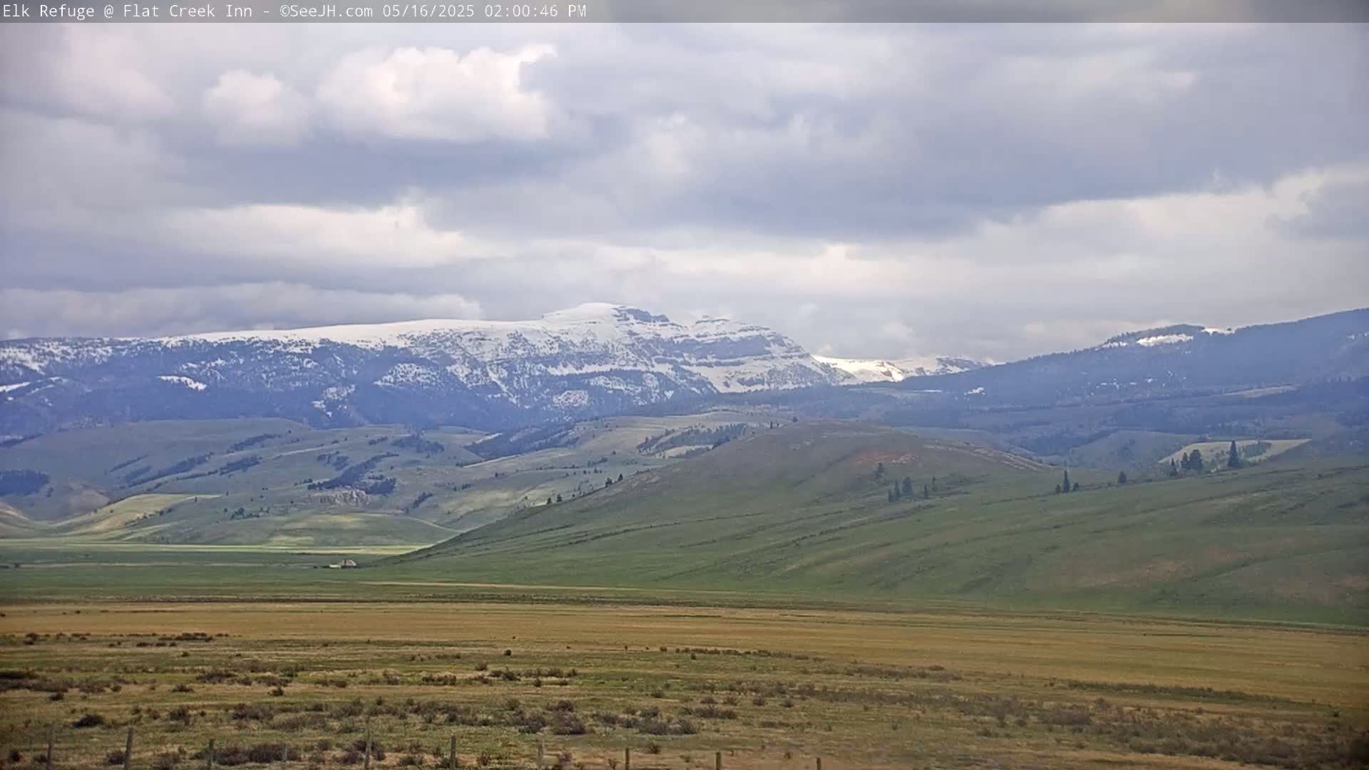 A mostly cloudy day reveals rolling green hills and distant mountains with patches of snow under a pale sky.