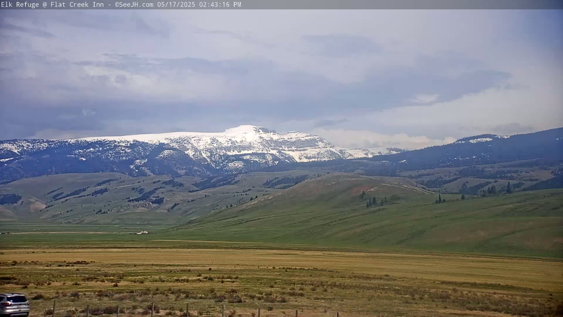 A partly cloudy day reveals snow-capped mountains in the distance, overlooking a vast expanse of gently rolling green and tan hills and plains.