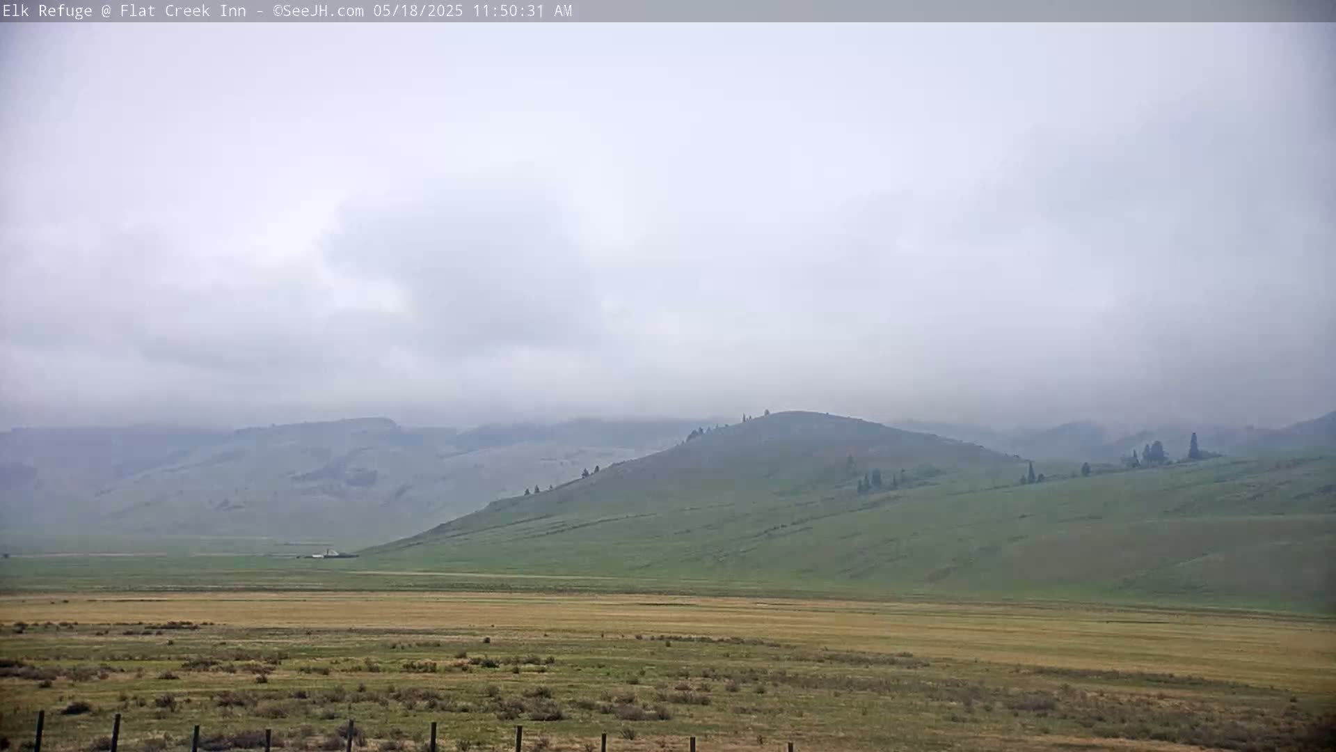 A hazy, overcast sky hangs over a wide, flat valley bordered by rolling green hills.