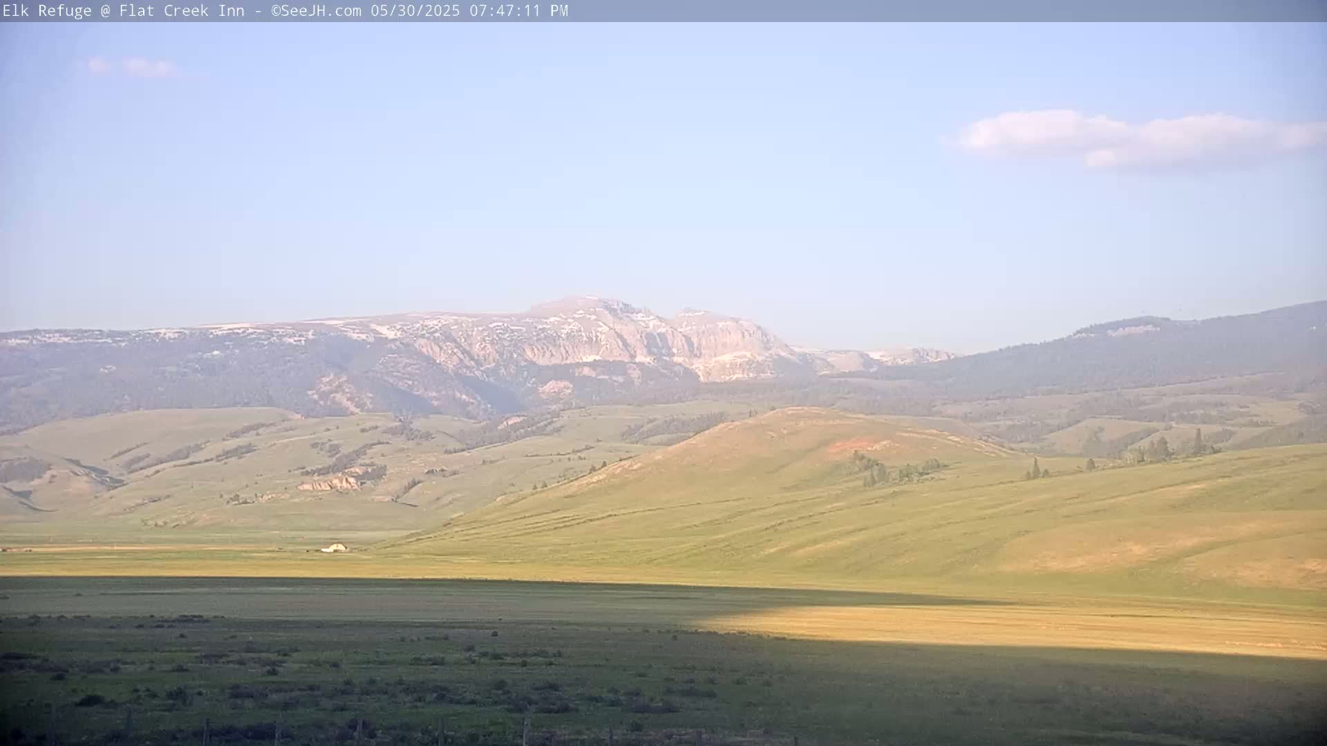 A mostly sunny day reveals rolling green hills and distant mountains with patches of snow under a pale blue sky.