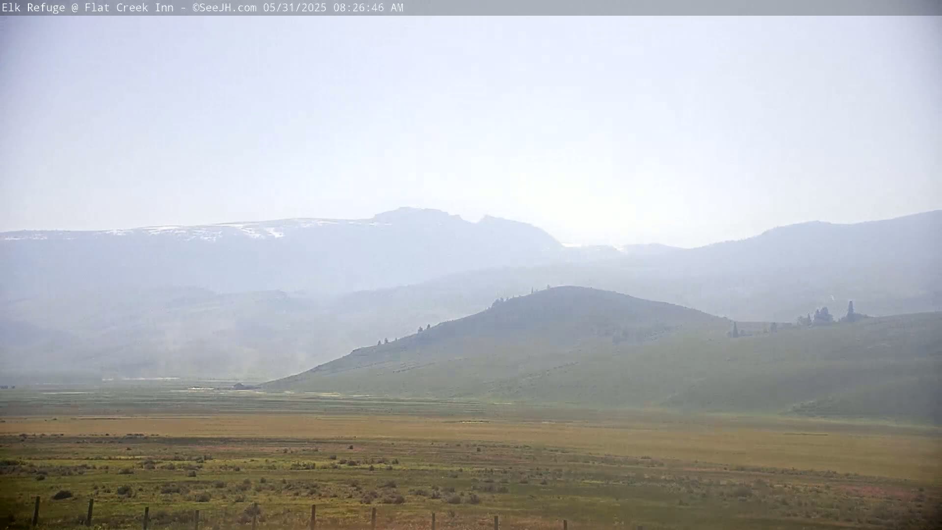 A hazy, flat valley stretches out before a range of distant, hazy mountains with patches of snow visible on the highest peaks.