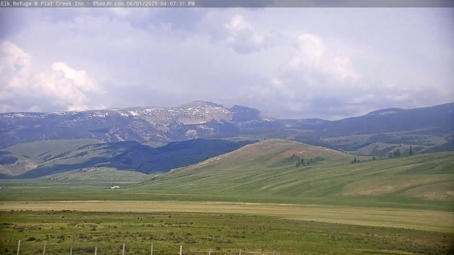 A partly cloudy day reveals rolling green hills and distant mountains with patches of snow under a mostly cloudy sky.