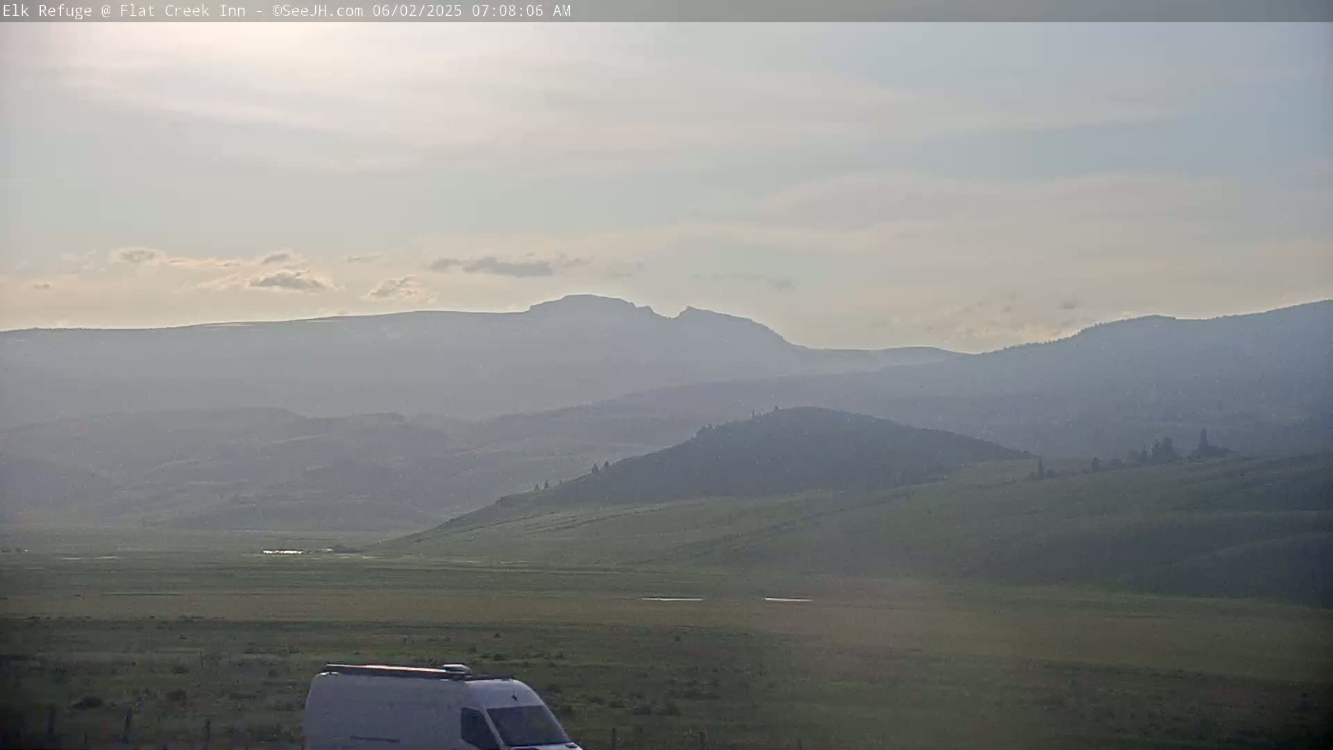 A hazy, light-colored sky overlooks a vast, gently rolling landscape of green hills and distant mountains, with a white van partially visible in the lower right corner.