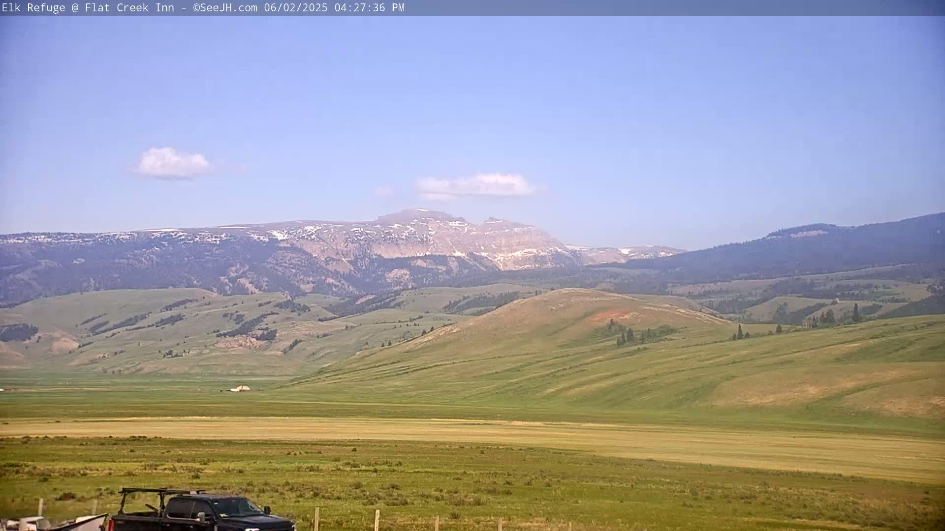 A mostly sunny day reveals a vast, rolling green landscape leading to distant mountains with patches of snow under a clear, light blue sky.