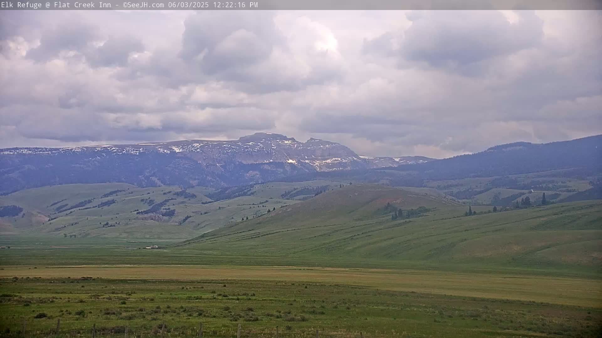 A wide, flat valley of green grasslands stretches to a range of mountains, some with patches of snow, under a cloudy sky.