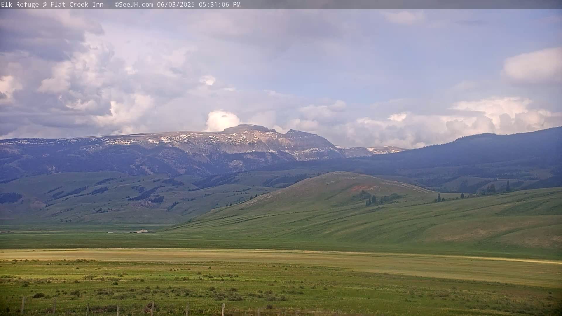 A wide, grassy valley stretches towards snow-capped mountains under a partly cloudy sky.