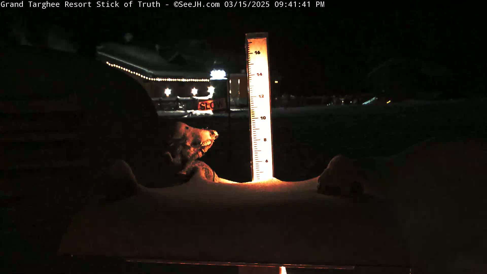 A snow-covered surface at night shows a illuminated snow depth gauge partially buried in the snow, with a blurry building and lights in the background.