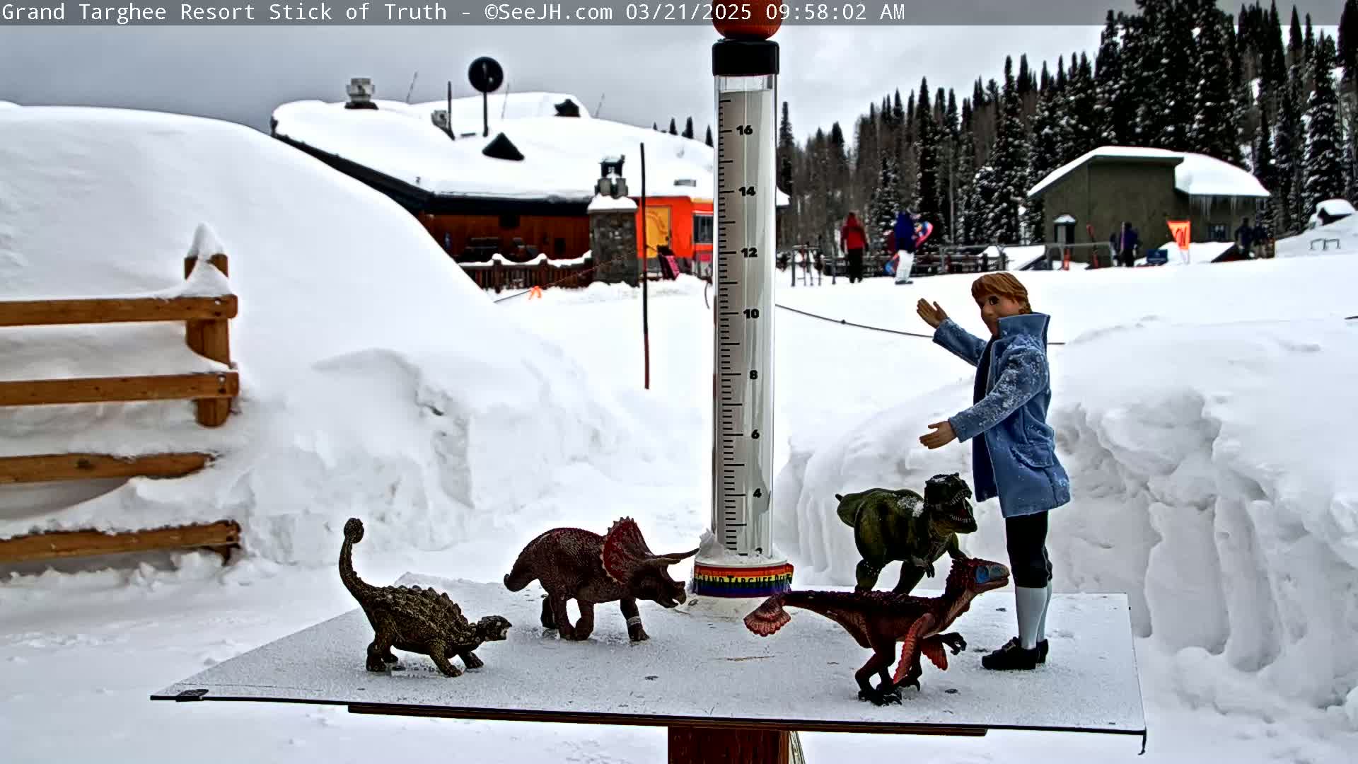 A snow-covered scene shows a doll standing near a snow gauge with several dinosaur figurines in front of a snowy building and a forest.