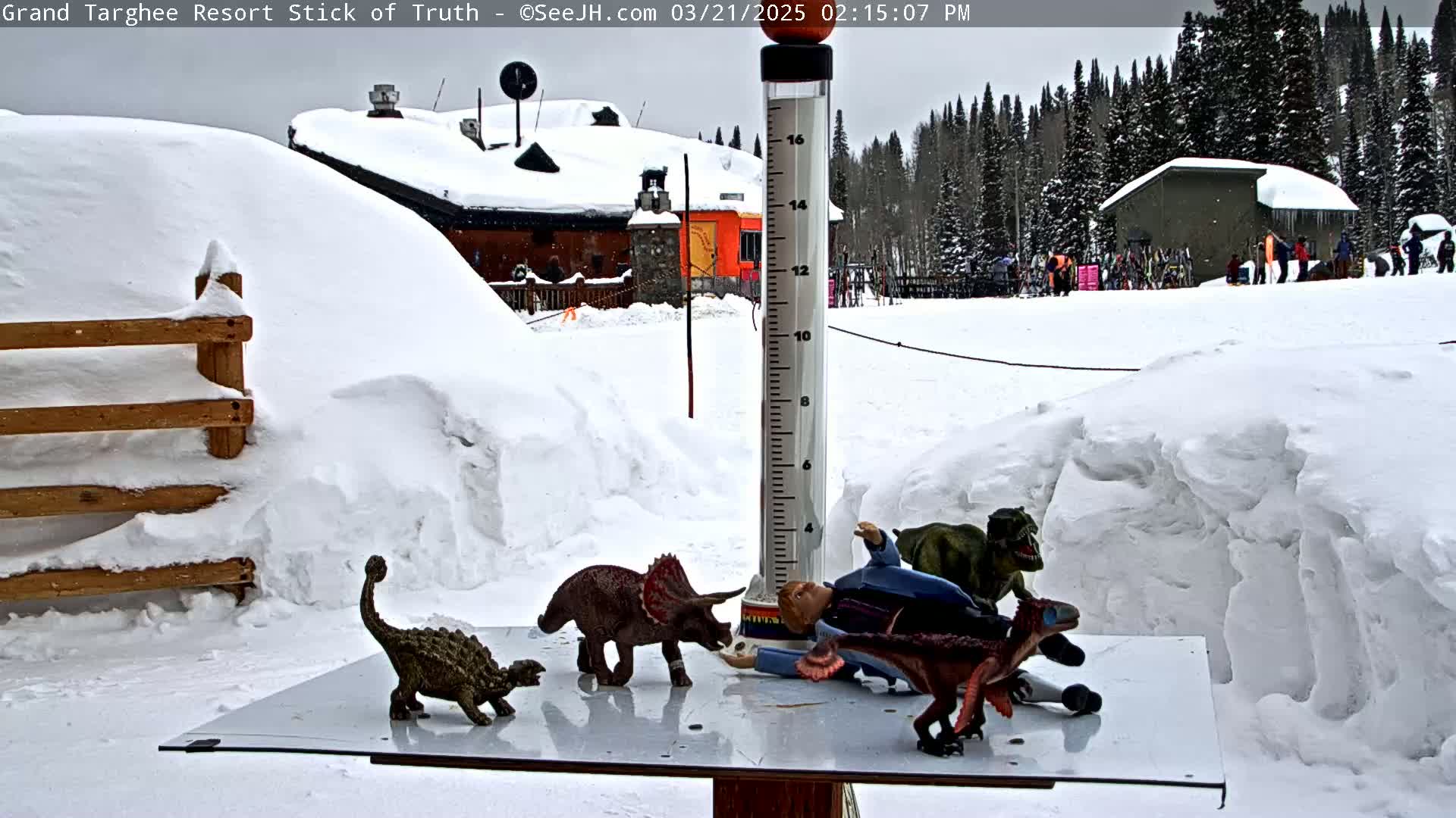 A snow-covered scene shows several dinosaur toys on a table near a snow-covered wooden fence and a building in the background, under an overcast sky.