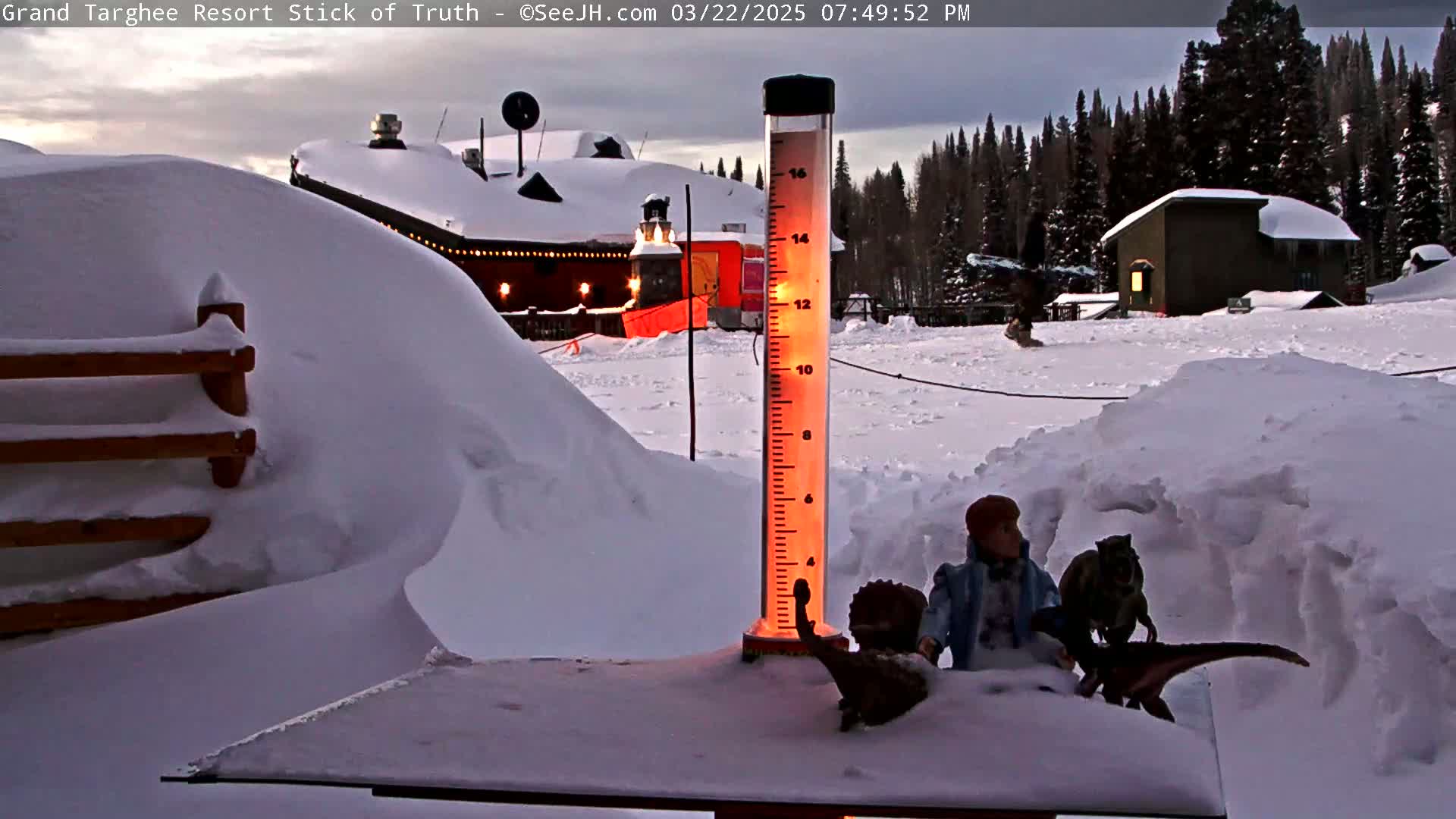 A snow-covered landscape at dusk features a lit rain gauge, snow drifts, a wooden fence, and buildings in the background.