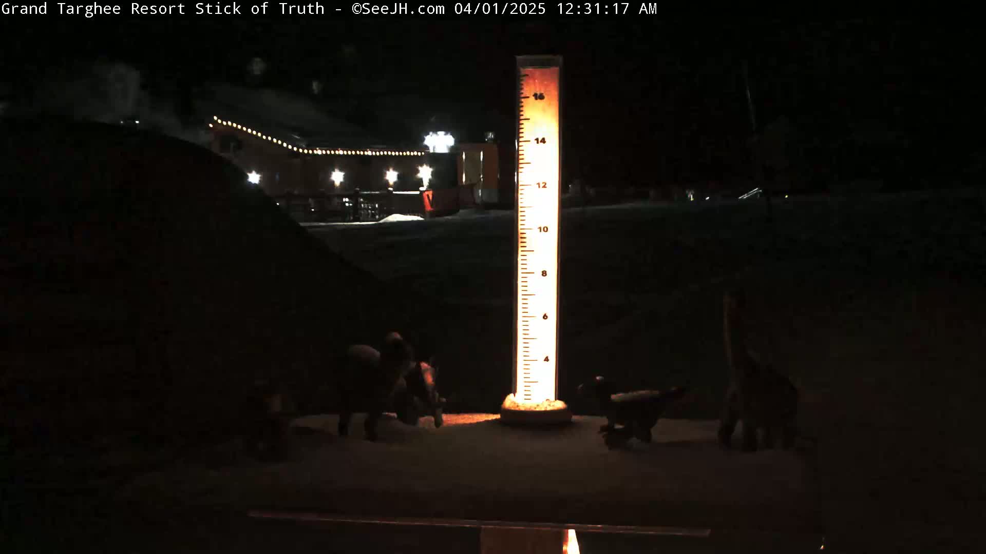 At night, a tall illuminated measuring stick stands in the snow near small dark figures, with a building with lights visible in the background.