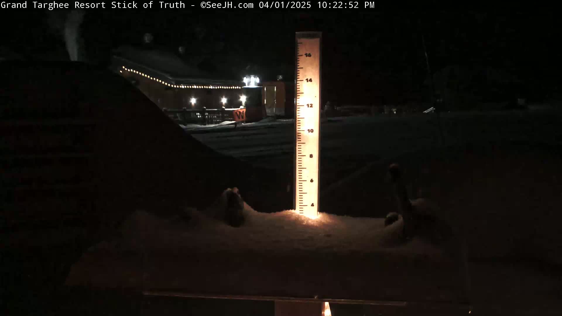 A snow-covered ground with a illuminated snow depth gauge in the foreground, showing a building with lights in the background at night.