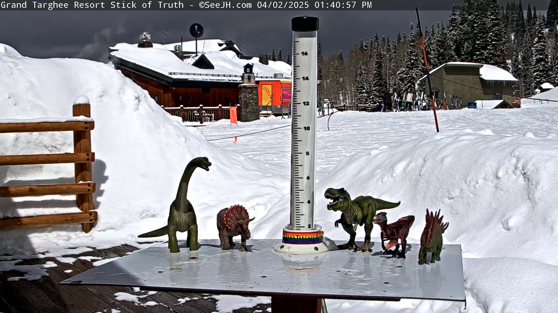A snow-covered outdoor scene shows a snow stick, several plastic dinosaur toys arranged on a white surface, and a snow-covered building in the background under a partly sunny sky.