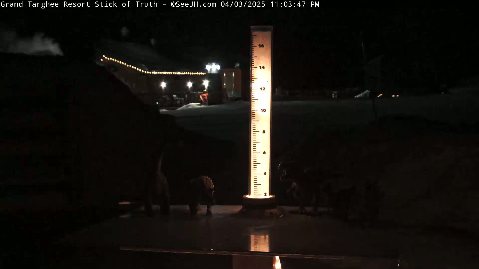 At night, a tall, illuminated measuring stick stands in a snow-covered area near a building with lights.