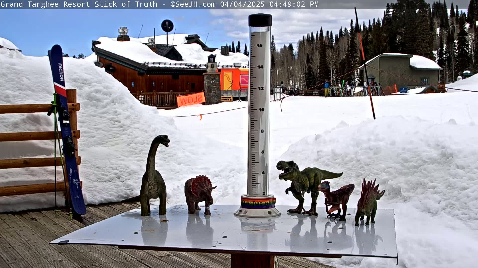 A snow-covered outdoor scene shows a wooden table with several plastic dinosaur toys arranged around a rain gauge, a pair of skis leaning against a wooden fence, and a building in the background under a partly cloudy sky.