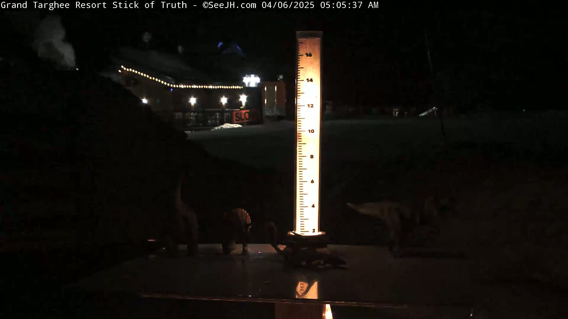At night, a tall, illuminated measuring device sits on a dark surface near a dimly lit building with string lights.