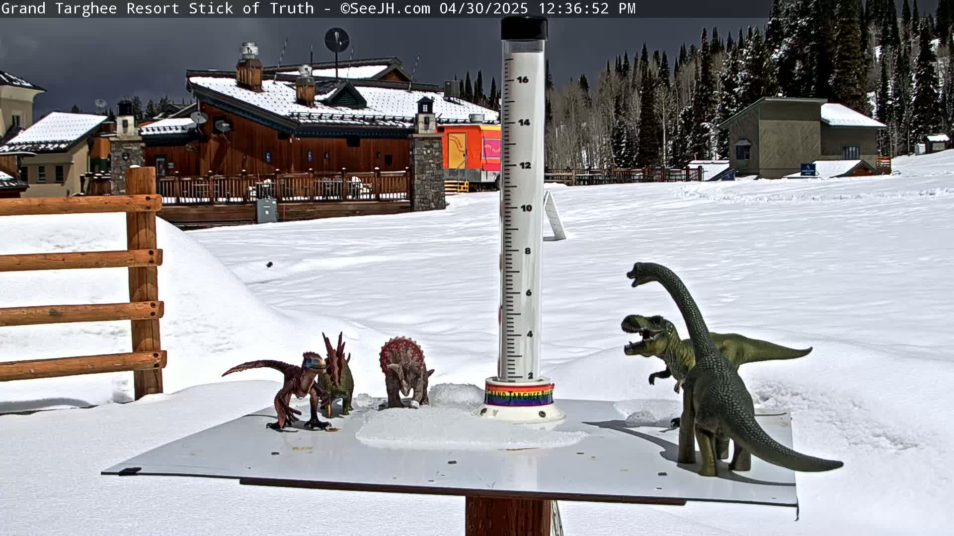 A snow-covered scene shows several dinosaur toys arranged on a table next to a tall measuring device, with buildings and a snow-covered forest in the background under a partly cloudy sky.