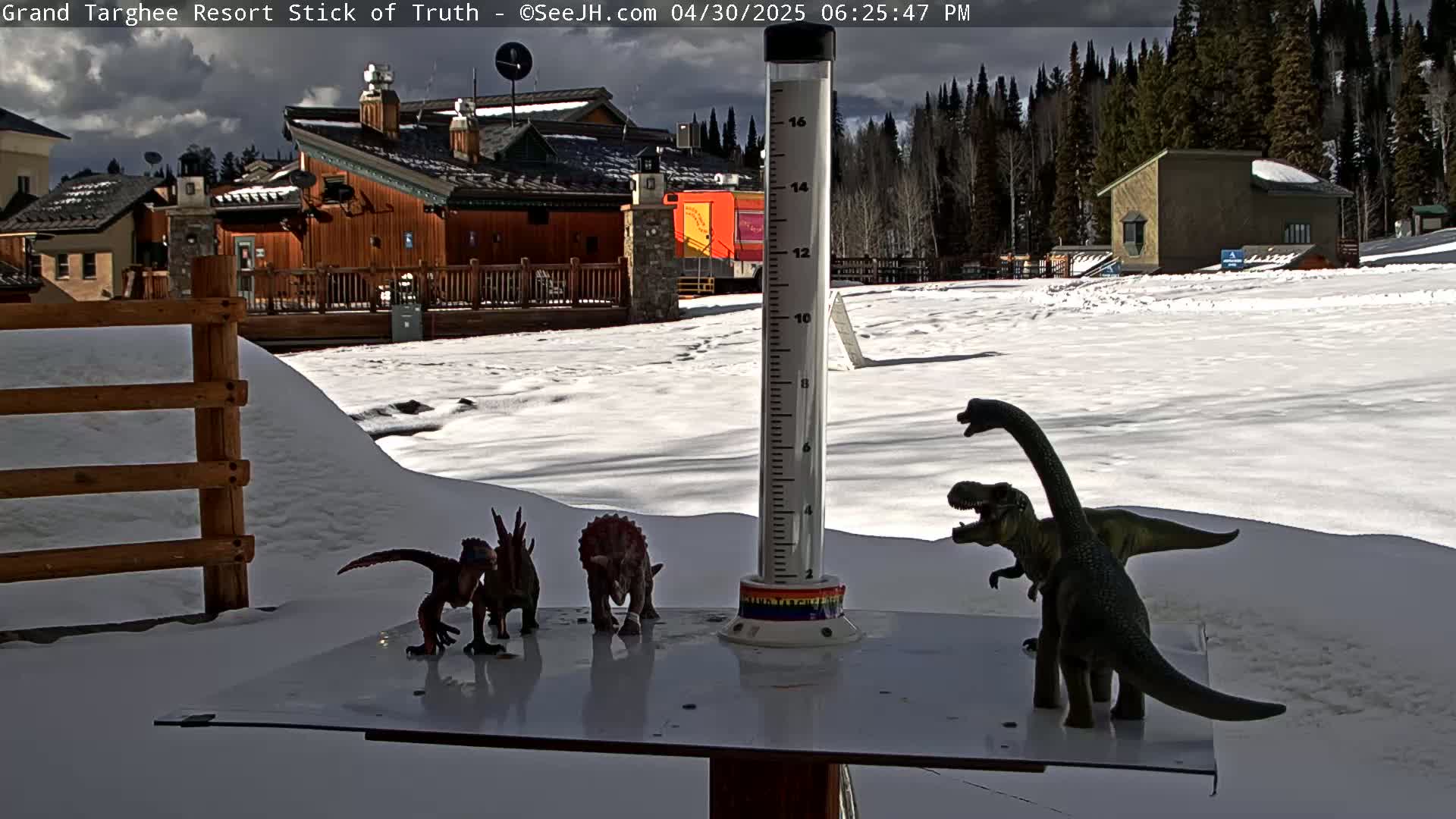 A snow-covered scene shows several toy dinosaurs arranged on a table near a weather gauge in front of a building complex and a snow-covered field with a forest in the background under a partly cloudy sky.